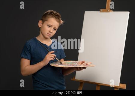 Hält eine Palette mit Farben kleinen Jungen in blauem T-Shirt traurig neben der Staffelei mit Leinwand auf sie stehen. School of Arts Konzept Stockfoto