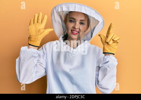 Schöne blonde kaukasische Frau trägt schützende Imker Uniform zeigen und Zeigt mit den Fingern Nummer sieben und lächelt zuversichtlich und Ha Stockfoto