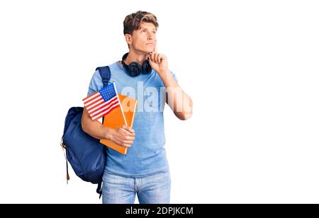 Junger gutaussehender Mann trägt Student Rucksack und Kopfhörer halten Buch und amerika Flagge ernstes Gesicht denken über Frage mit Hand auf Kinn, thoug Stockfoto