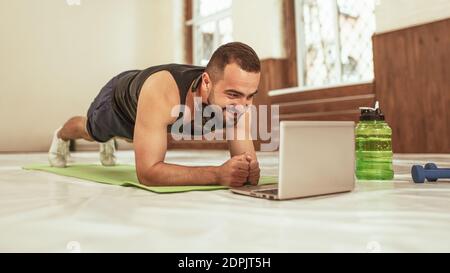 Fitnesstrainer, der während der Selbstisolation mit einem Kunden eine Online-Videokonferenz mit einem Laptop auf der Matte macht. Junger Mann, der etwas Besonderes macht Stockfoto