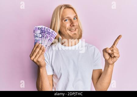 Kaukasischer junger Mann mit langen Haaren, der 100 philippinische Peso hält Banknoten lächeln glücklich zeigen mit Hand und Finger auf die Seite Stockfoto