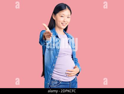 Junge schöne chinesische Frau schwanger erwartet Baby lächelnd freundlich bietet Handschlag als Gruß und einladend. Erfolgreiches Geschäft. Stockfoto