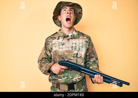 Junger gutaussehender Mann trägt Tarnuniform Armee hält Schrotflinte wütend und verrückt schreien frustriert und wütend, schreien mit Wut nach oben. Stockfoto