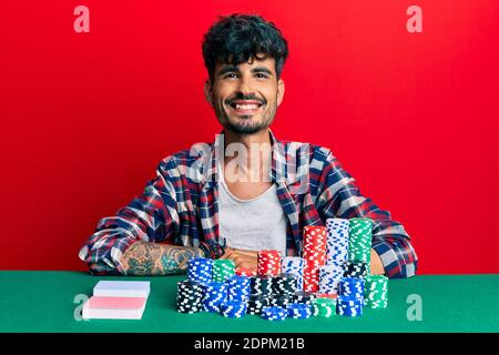 Junge hispanic Mann sitzt auf dem Tisch mit Poker-Chips und Karten mit einem glücklichen und kühlen Lächeln auf dem Gesicht. Glückliche Person. Stockfoto