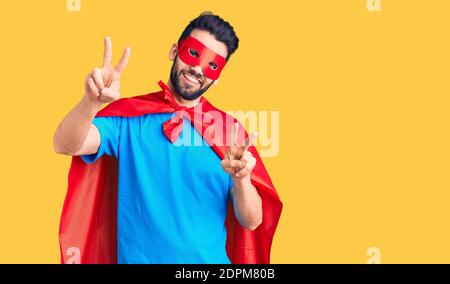 Junger schöner Mann mit Bart trägt Superhelden Kostüm lächelnd Blick auf die Kamera zeigt Finger tun Siegeszeichen. Nummer zwei. Stockfoto