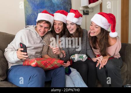 Vier Geschwister im College-Alter tragen Weihnachtsmützen Blick auf Selfies auf Telefon Stockfoto