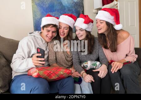 Vier Geschwister im College-Alter tragen Weihnachtsmützen Blick auf Selfies auf Telefon Stockfoto