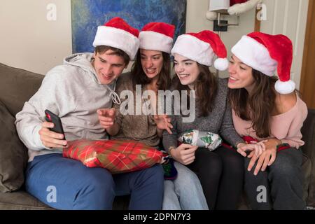 Vier Geschwister im College-Alter tragen Weihnachtsmützen Blick auf Selfies auf Telefon Stockfoto