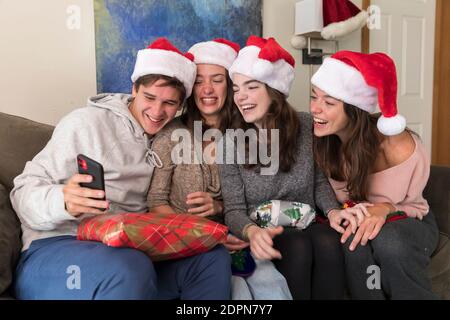 Vier Geschwister im College-Alter tragen Weihnachtsmützen Blick auf Selfies auf Telefon Stockfoto