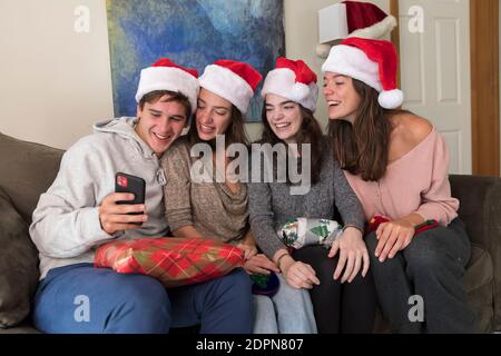 Vier Geschwister im College-Alter tragen Weihnachtsmützen Blick auf Selfies auf Telefon Stockfoto