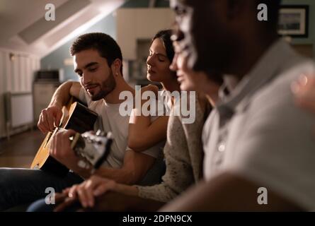 Gruppe junger multiethnischer Menschen, die jungen männlichen Musikers zuhören Spielen akustische Gitarre während der Freunde treffen zu Hause Stockfoto