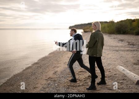 Ganzer Körper glücklich junges Paar werfen Kieselsteine in See Wasser Während Sie Zeit in der abendlichen Landschaft zusammen verbringen Stockfoto