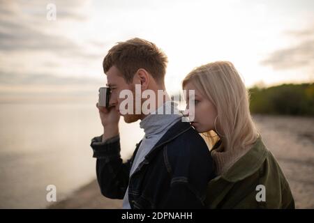 Seitenansicht der blonden jungen Frau hinter Freund mit stehen Fotokamera auf verschwommenem Hintergrund des Seeufers im Herbst Auf dem Land Stockfoto