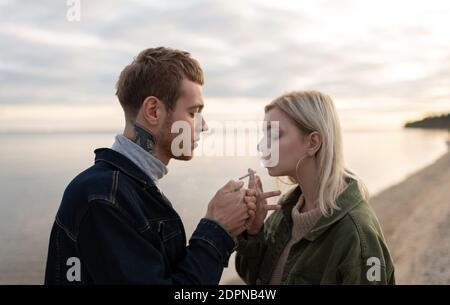 Seitenansicht von jungen Hipstern, die im Stehen Gras rauchen Gegen ruhigen See und bewölkten Himmel während des Datums im Herbst Auf dem Land Stockfoto