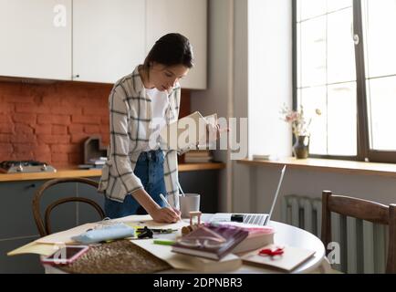 Fokussierte junge Frau im Hemd stehend am Tisch in der Küche Und Notizen aus dem Lehrbuch zu schreiben, während Sie zu Hause studieren Stockfoto