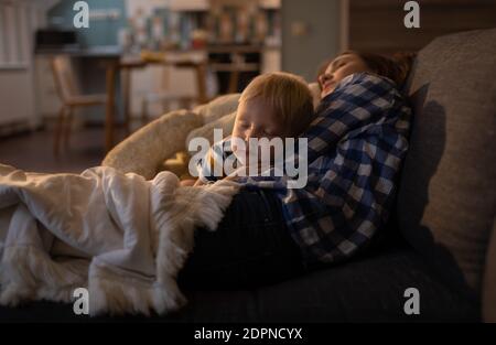 Ruhiges blondes kleines männliches Kind, das in den Armen einschläft Von jungen Schwester in Freizeitkleidung auf bequemen Sofa an Wohnzimmer Stockfoto