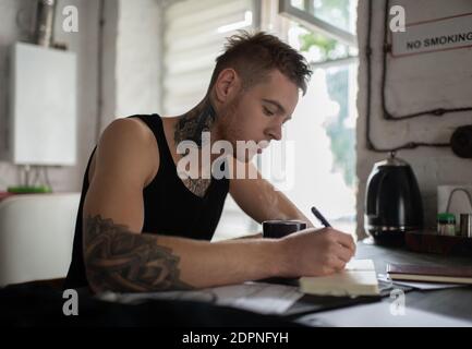 Seitenansicht des inspirierten Schriftstellers in schwarzem T-Shirt schreiben von Geben Sie ein Notizbuch und trinken Sie morgens Kaffee am Tisch Stockfoto