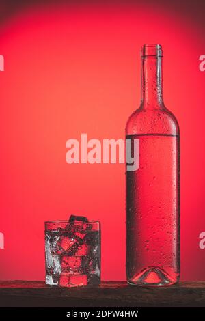 Erfrischendes kaltes Getränk in der Flasche mit Tropfen und Glas mit Eis auf rotem Hintergrund. Erfrischende Eisgetränke Konzept. Stockfoto