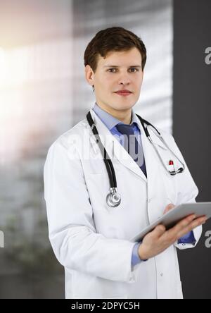 Ein junger, freundlicher Arzt überprüft einige Informationen auf seinem Computer-Tablet. Porträt eines Facharztes bei der Arbeit in der sonnigen Klinik Stockfoto