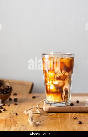 Kalter erfrischender Kaffee mit Eis und cremigen Wolken in einem Glas auf einem Holzbrett Nahaufnahme, Kakaobohnen in Kokosnussschale Stockfoto