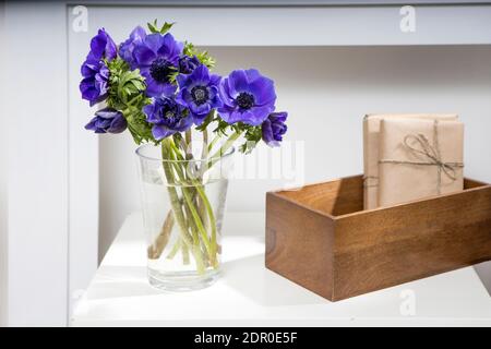 Bouquet der blauen Anemonen in einer Glasvase auf einem weißen Tisch nahe der hellgrauen Wand. Die Holzkiste mit zwei eingepackten Geschenk. Vertikaler Rahmen. Kopieren Sie den SPAC Stockfoto