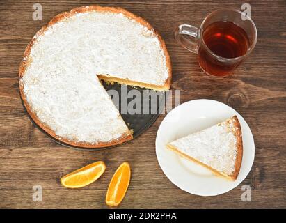 Stück hausgemachter Kuchen mit Zitronen- und Orangenfüllung, bedeckt mit Puderzucker auf einem Holztisch in der Nähe von Orangenfrüchten und einer Tasse Tee Stockfoto