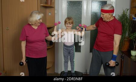 Enkelin und ältere Großeltern in Sportbekleidung machen Sport Workout Hanteln Übungen zu Hause. Kaukasischen Senior paar Großmutter Großvater mit Kind Kind Mädchen tun Fitness Gewichtheben Stockfoto