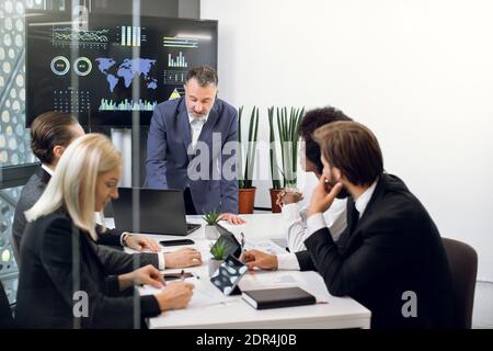 Reifer männlicher Chef lehnt sich an den Tisch und spricht mit seinem Team, professionellen erfolgreichen multiethnischen männlichen und weiblichen Kollegen, über die Zusammenarbeit Stockfoto