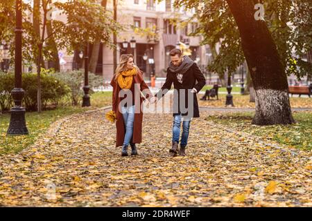 Pärchen, die im Park herumlaufen und Hände halten. Außenaufnahme eines verliebten jungen Paares, das auf einem Pfad durch einen Herbstpark geht. Herbstfarbiges Bild Stockfoto
