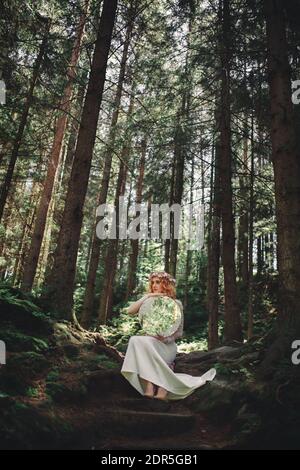 Schöne junge rothaarige Braut im Wald mit einem Blumenkranz auf dem Kopf und Spiegel. Frau in langen weißen Kleid im Freien am Sommertag. Hochzeit Stockfoto