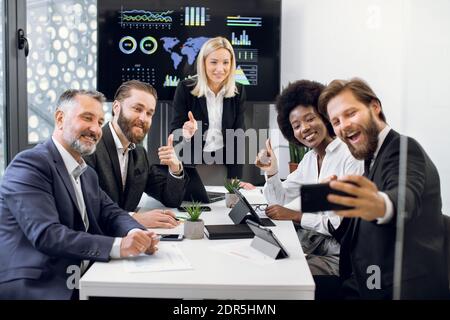 Fröhliches internationales kreatives, vielfältiges Business-Team in formeller Kleidung, Selfie und Daumen nach oben, am Tisch im Büro sitzend. Unternehmen Stockfoto