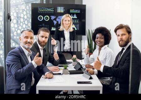 Team von professionellen hochqualifizierten multiethnischen Team von Männern und Frauen, sitzen am Tisch im Bürozimmer mit großen Wand-Bildschirm, und zeigen Stockfoto