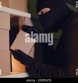 Ein Dieb in einer schwarzen Haube stiehlt im Dunkeln ein Paket in einem Lagerhaus. Konzept der Probleme mit Diebstahl von Waren und Postpaketen Stockfoto