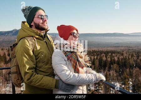 Seitenansicht von fröhlichen jungen Dates in Winterbekleidung und Sonnenbrillen Blick nach vorne gegen blauen Himmel über schneebedeckten Bergen Stockfoto