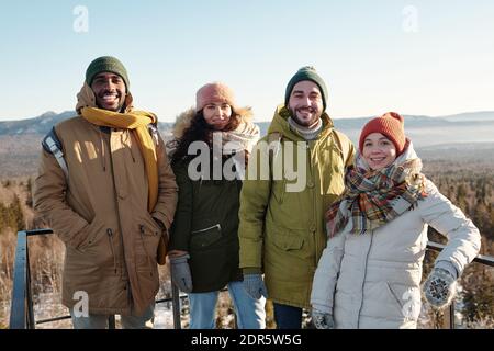 Fröhliche junge interkulturelle Freunde in Winterkleidung stehen vor Kamera gegen blauen Himmel über schneebedeckten Bergen während Entspannen Stockfoto