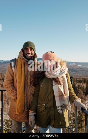 Fröhliche junge interkulturelle Dates in Wintermode stehen vor Kamera gegen blauen Himmel über Bergen mit Schnee bedeckt während Winterkälter Stockfoto