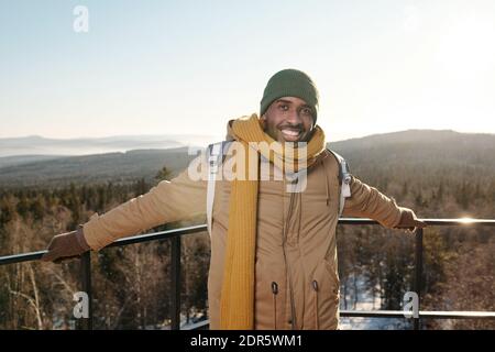 Glücklicher junger afrikanischer Mann in Winterkleidung, der vor ihm steht Kamera gegen klaren Himmel über Bergen und Mischwald auf Frostiger Wintertag Stockfoto