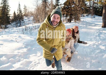 Glücklicher junger Mann in Winterkleidung, der zwischen Schneeverwehungen auf der Straße läuft Beim Ziehen Schlitten mit zwei fröhlichen Mädchen und Spaß haben Am Wintertag Stockfoto