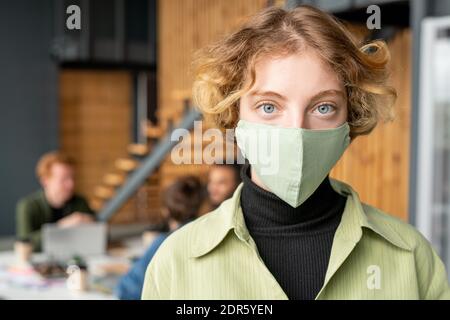 Junge blonde Büroangestellte in Casualwear und Schutzmaske Sie betrachten, während Sie vor der Kamera gegen stehen Kollegen Stockfoto