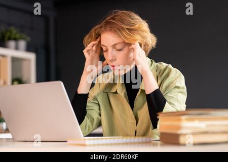 Junge blonde weibliche Software-Entwickler sitzen am Schreibtisch und versuchen Konzentrieren, während Sie den Kopf vor dem Laptop berühren Netzwerk Stockfoto