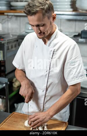 Junger männlicher Koch des modernen Restaurants in weißer Uniform hacken Pilze für Gemüse garnieren, während Sie am Tisch in der stehen Küche Stockfoto