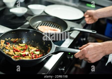 Hände des männlichen Küchenchefs rösten Stück Lachs und Gemüse Auf heißen Bratpfannen anbraten, während man am Elektroherd steht Und Kochen gesundes Mittagessen Stockfoto