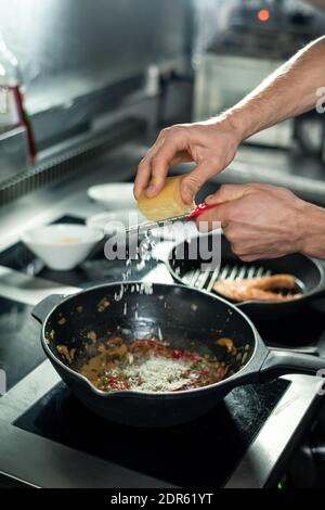 Hände des jungen professionellen Küchenchefs, der Käse in die Bratpfanne reibert Mit gehacktem Gemüse beim Kochen Eintopf für Kunden in der Küche Stockfoto