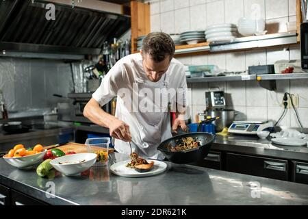 Junger männlicher Koch in Uniform hält Bratpfanne mit Gemüse Beim Bücken über den Tisch mit dem Teller eintoben und Essen geben Auf einem Stück gebratenen Lachs Stockfoto