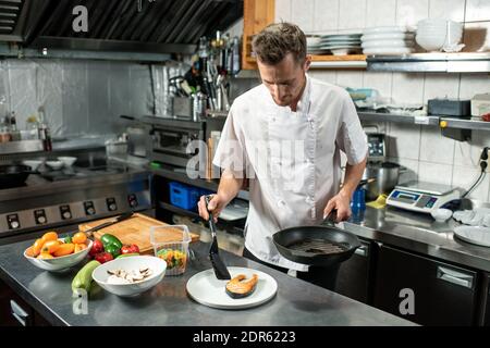 Moderner Chefkoch, der ein Stück gebratenen Lachs auf den Teller legt Beim Stehen an einem großen Küchentisch und beim Zubereiten des Essens Kunde Stockfoto