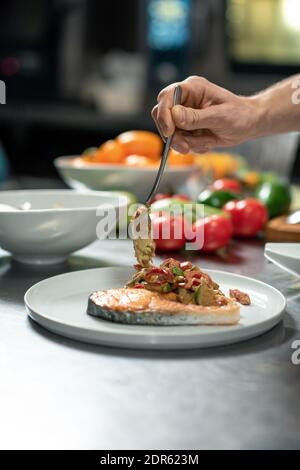 Hand des männlichen Küchenchefs, der heißen, appetitlichen Gemüseeintopf anlegt Teller mit einem Stück gebratenen Lachs während der Zubereitung einer leckeren Mahlzeit Für Restaurantkunden Stockfoto