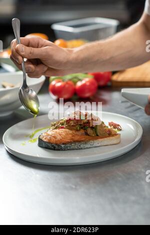 Hand des männlichen Chefkochs Zugabe von Olivenöl zu appetitlichen Gemüse Während der Zubereitung auf dem Teller mit einem Stück gebratenem Lachs eintoben Leckeres Essen für den Gast Stockfoto