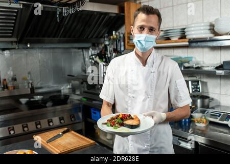 Junger männlicher Koch in weißer Uniform und Schutzmaske hält Teller mit gebratenem Lachs und Gemüse, während Sie davor stehen Der Kamera Stockfoto