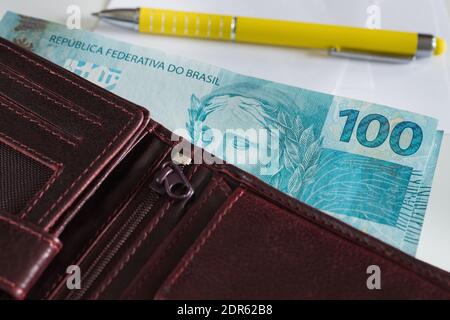 Brasilianische Banknoten, einhundert Reais ragen aus der Brieftasche, ein Stück Papier mit einem Stift daneben Stockfoto