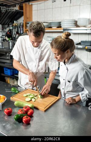 Junger professioneller Chefkoch, der seiner Auszubildenden zeigt, wie es geht Kochen Sie frische Zucchini, während beide am Tisch in der stehen Küche des Restaurants Stockfoto